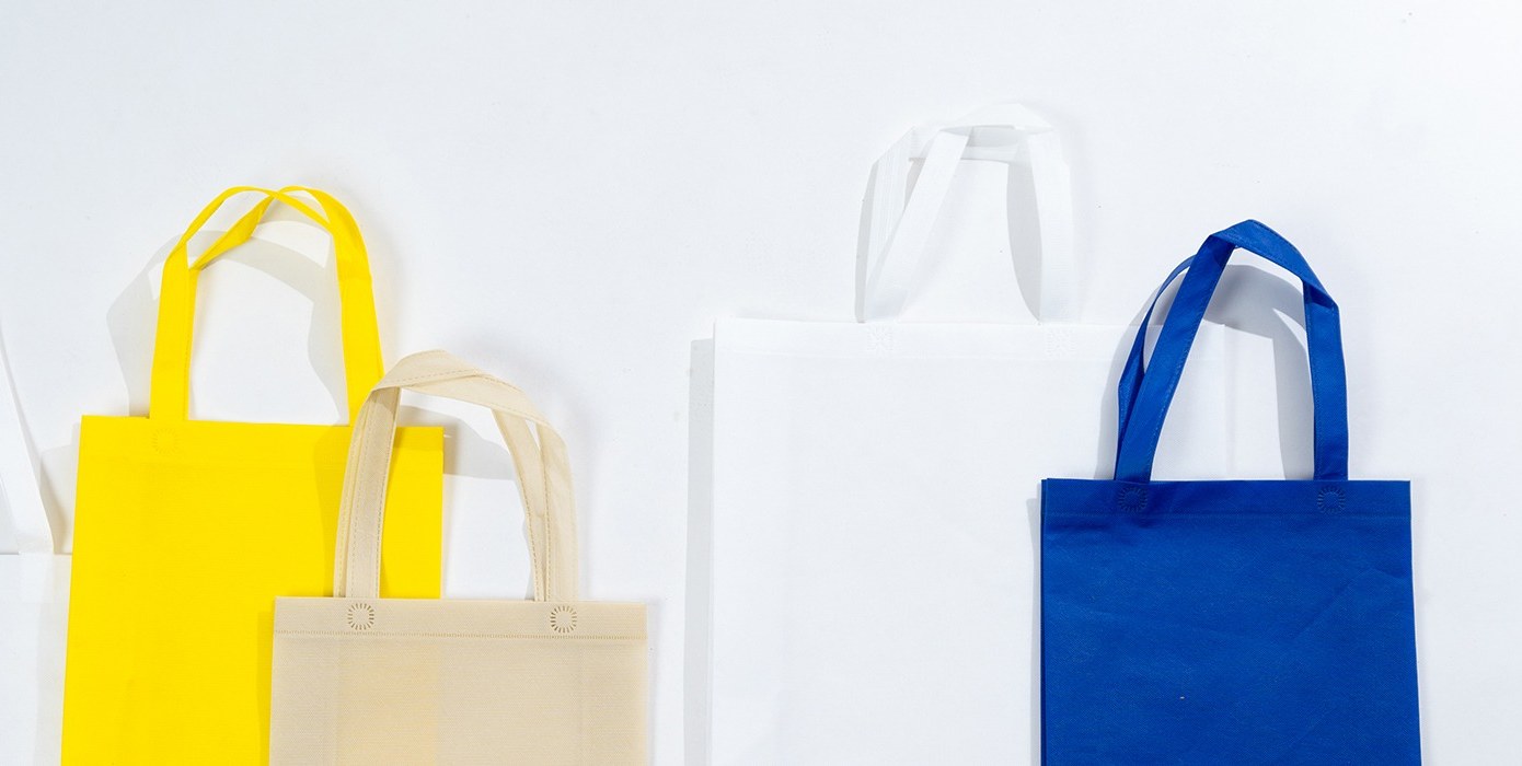 Five plain non woven tote bags in white, yellow, cream and blue, standing upright